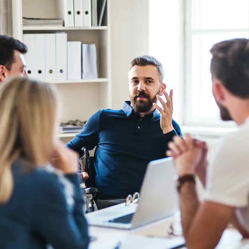 Innovation-team-testimonial Group of staff sitting around a desk, One man is talking with expressive hand gusture