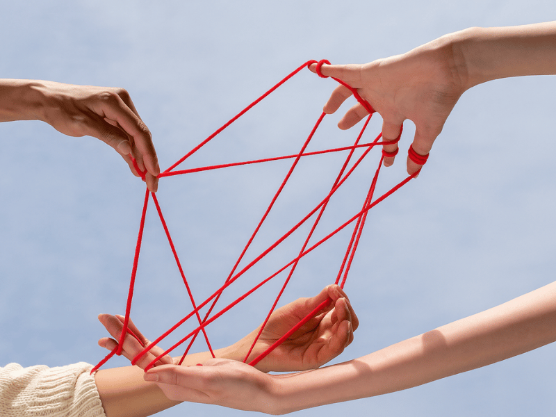 2 pairs of hands holding red string - the red string connects the peoples fingers against blue sky as a metaphor for co-regulation